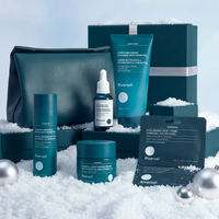 Riverol skincare gift set arranged in a snowy display with teal and black packaging, including bottles, jars, a pouch, and boxes, alongside silver ornaments against a soft blue sky backdrop.