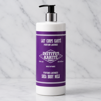A white bottle of Institut Karite Paris Shea Body Milk with a purple label and black pump dispenser, set on a white marble surface against a marble background.