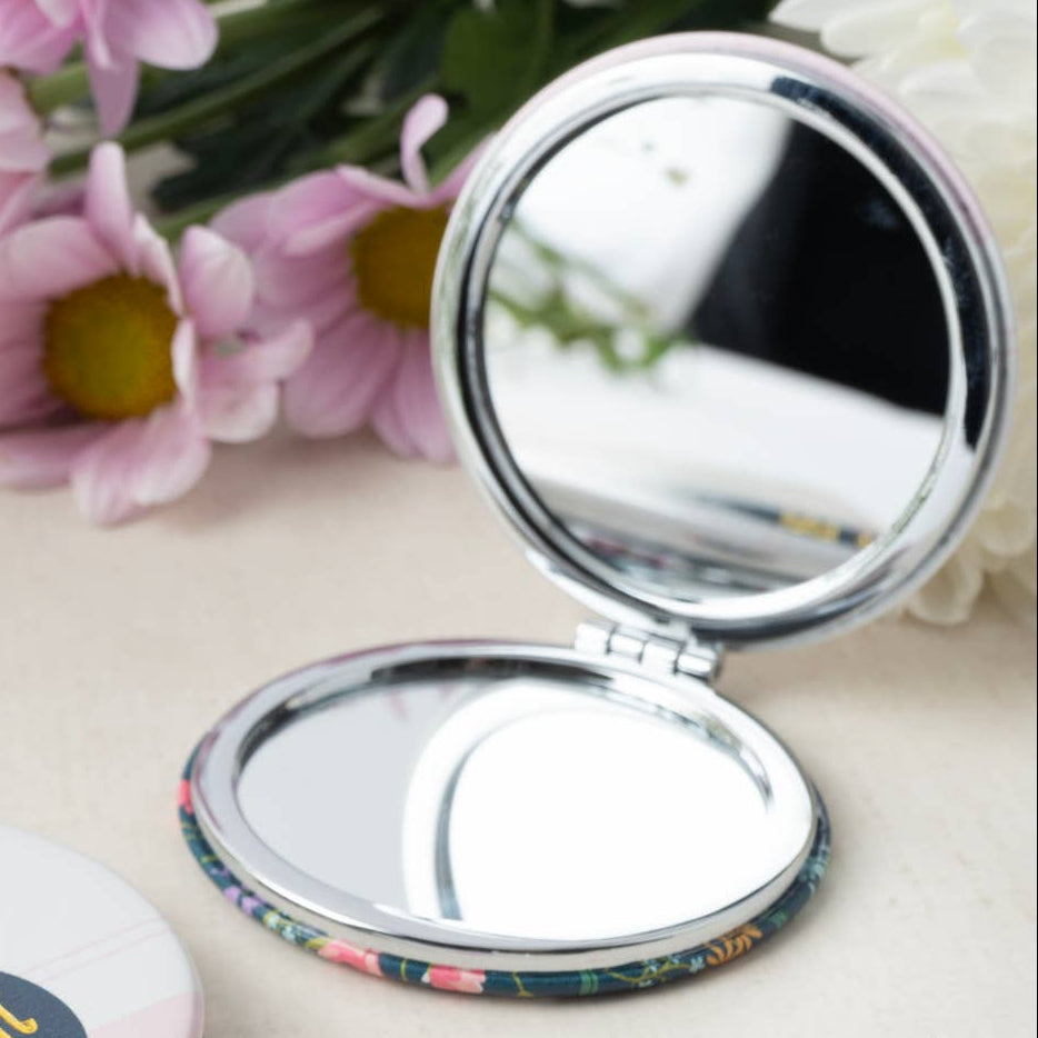 Two round compact mirrors on a light beige surface with pink flowers in the background. One compact is closed, showing a floral lid with the text 'BLOOM! fabulous' in gold on a dark blue label.