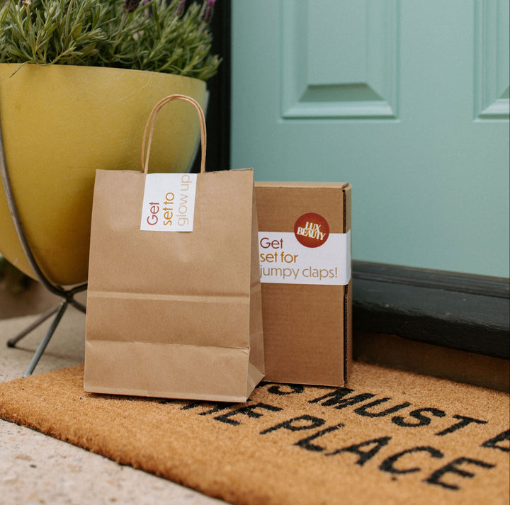 Brown paper shopping bag with a LUXBEAUTY.COM label on a doormat in front of a turquoise door.