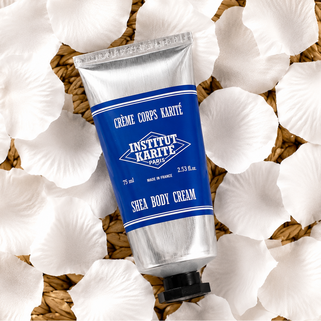A silver squeeze tube of Institut Karité Paris Shea Body Cream with a blue label, styled on a bed of white flower petals over a woven straw mat.