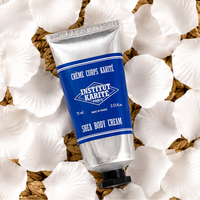 A silver squeeze tube of Institut Karité Paris Shea Body Cream with a blue label, styled on a bed of white flower petals over a woven straw mat.