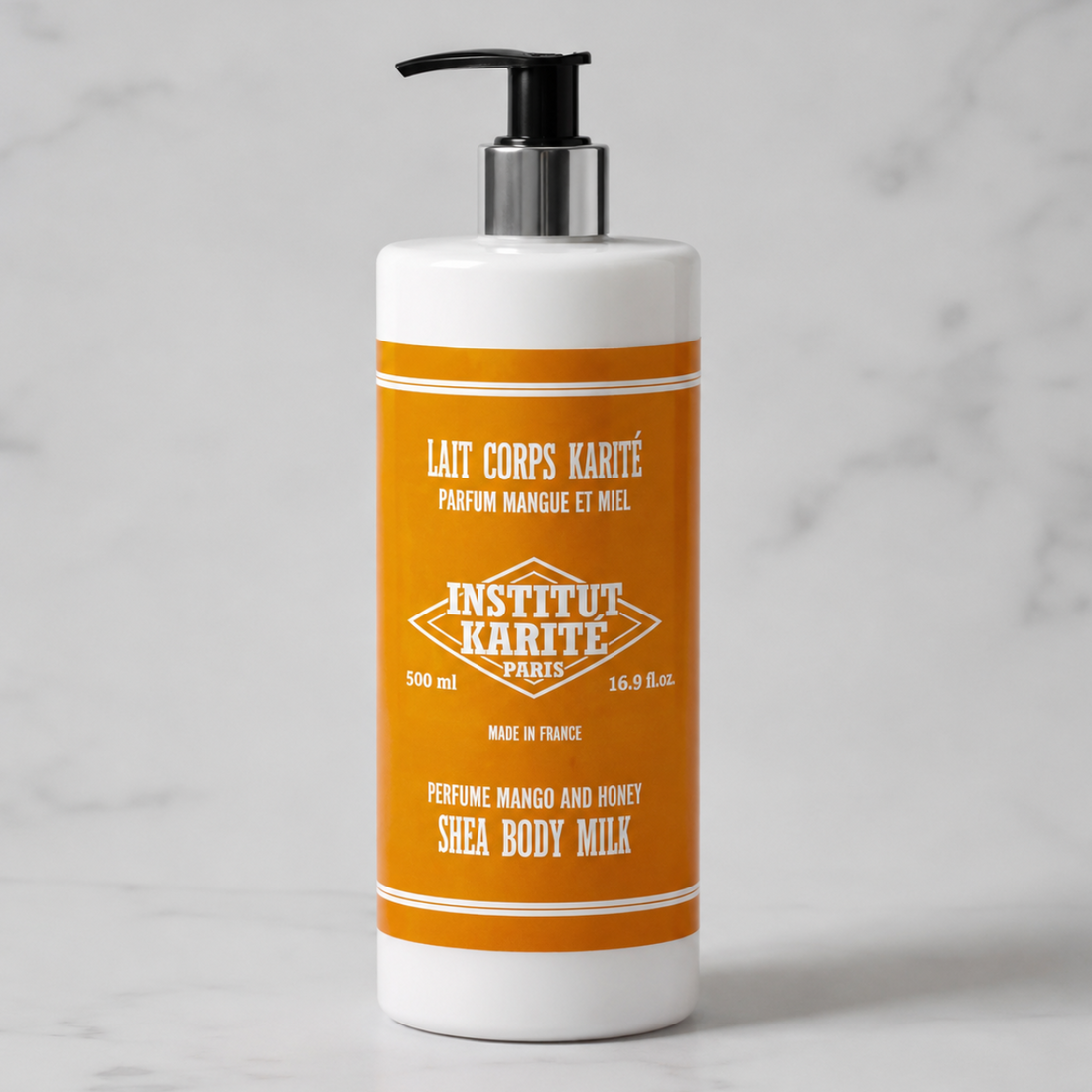A white plastic pump bottle of Institut Karité Paris Shea Body Milk with a black pump and a prominent orange label, set against a light gray marble background.