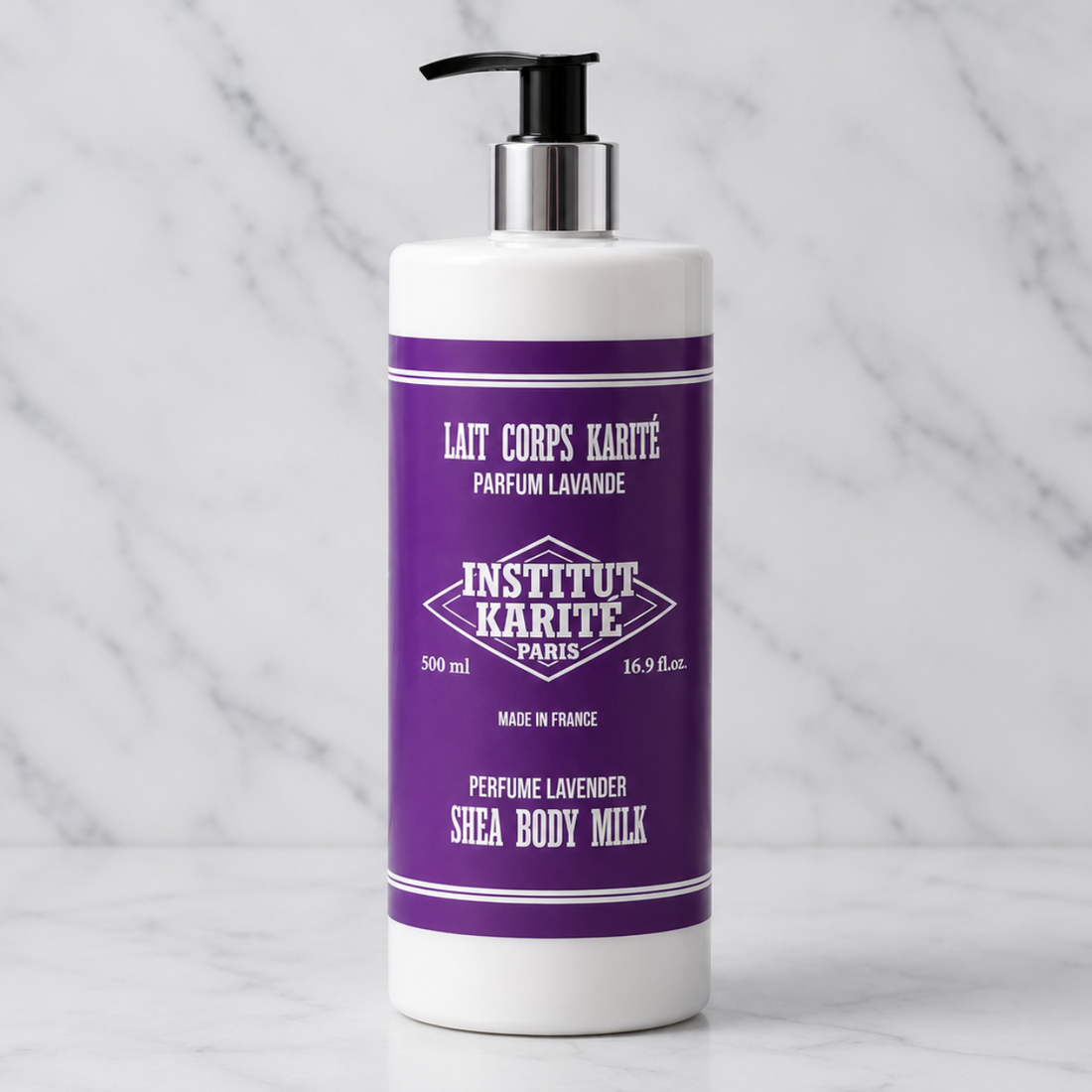 A white bottle of Institut Karite Paris Shea Body Milk with a purple label and black pump dispenser, set on a white marble surface against a marble background.
