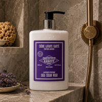 A 500 ml bottle of Institut Karitte Lavender Perfume Shea Cream Wash sits on a stone shower ledge in a bathroom setting. The bottle is white with a purple label and a black pump dispenser, set against a background of gray stone tiles and a brass shower fixture.