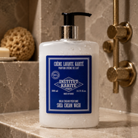A bottle of Institut Karité Paris Shea Cream Wash with a black pump and blue label, set on a beige marble bathroom counter with a gold shower fixture in the background.